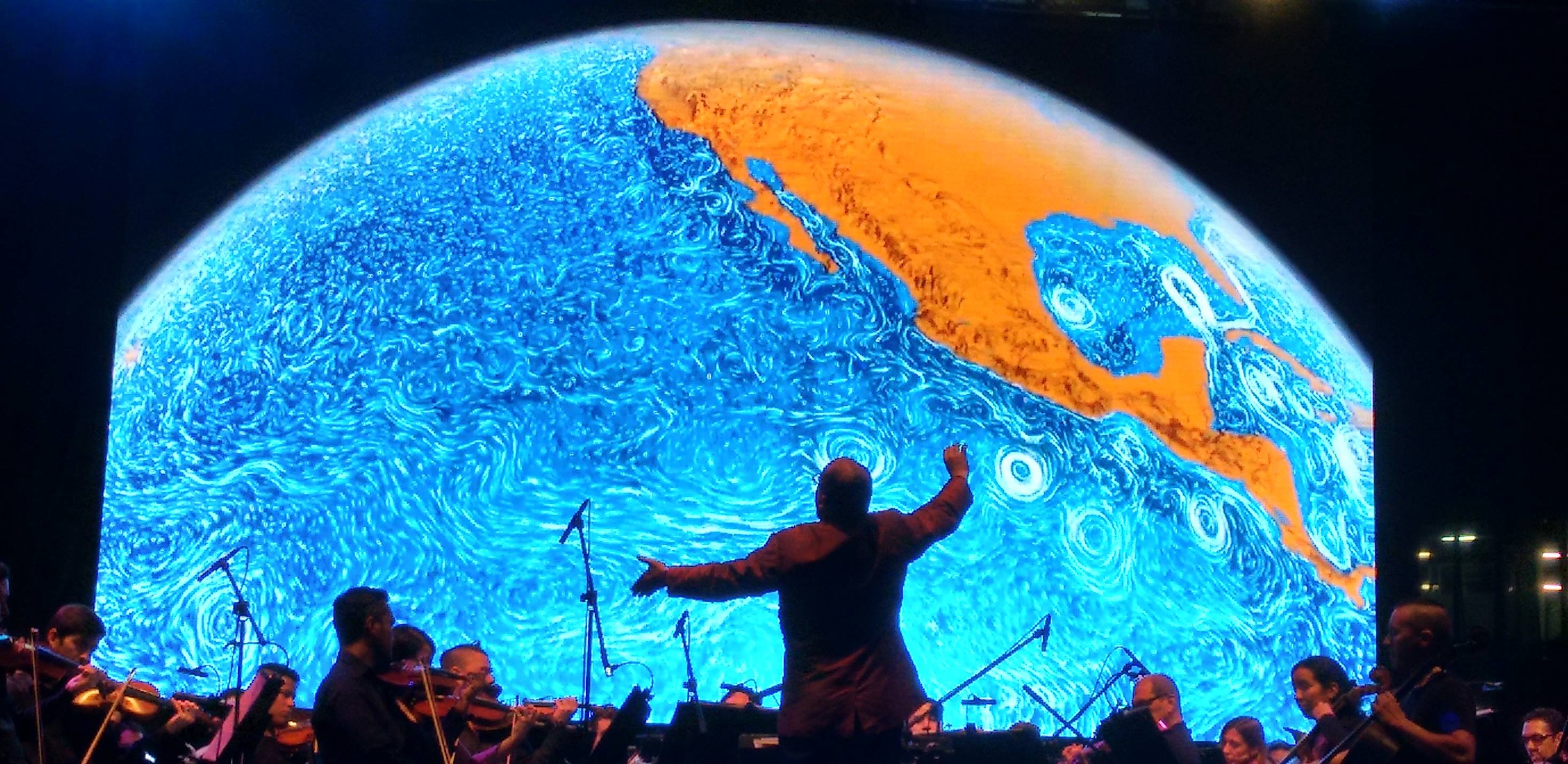 Fotografía. En el centro, un director de orquesta levanta las manos, de espaldas al lector. A su alrededor hay músicostocando violines y violonchelos. Al fondo, muy grande, se ve la imagen del planeta tierra, una parte del mar con lineas azules que representan las corrientes y una parte del continente, color café